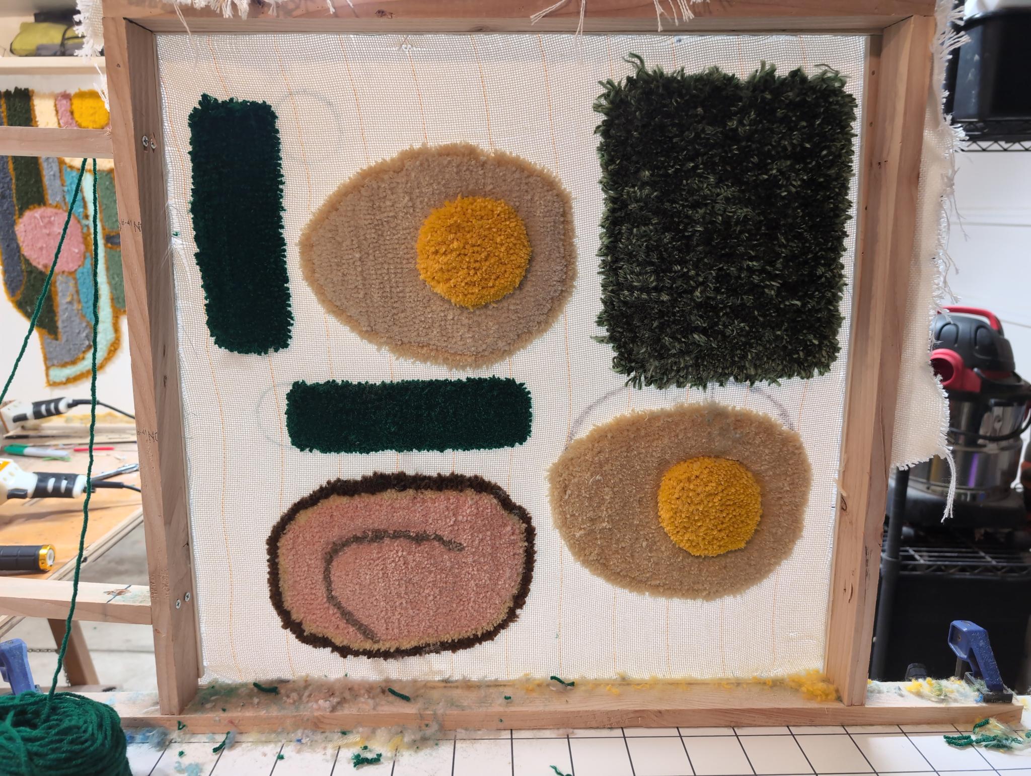 Tufted ramen ingredients in progress on the stretcher frame: two egg discs, pink chashu, dark green nori, and green onion segments