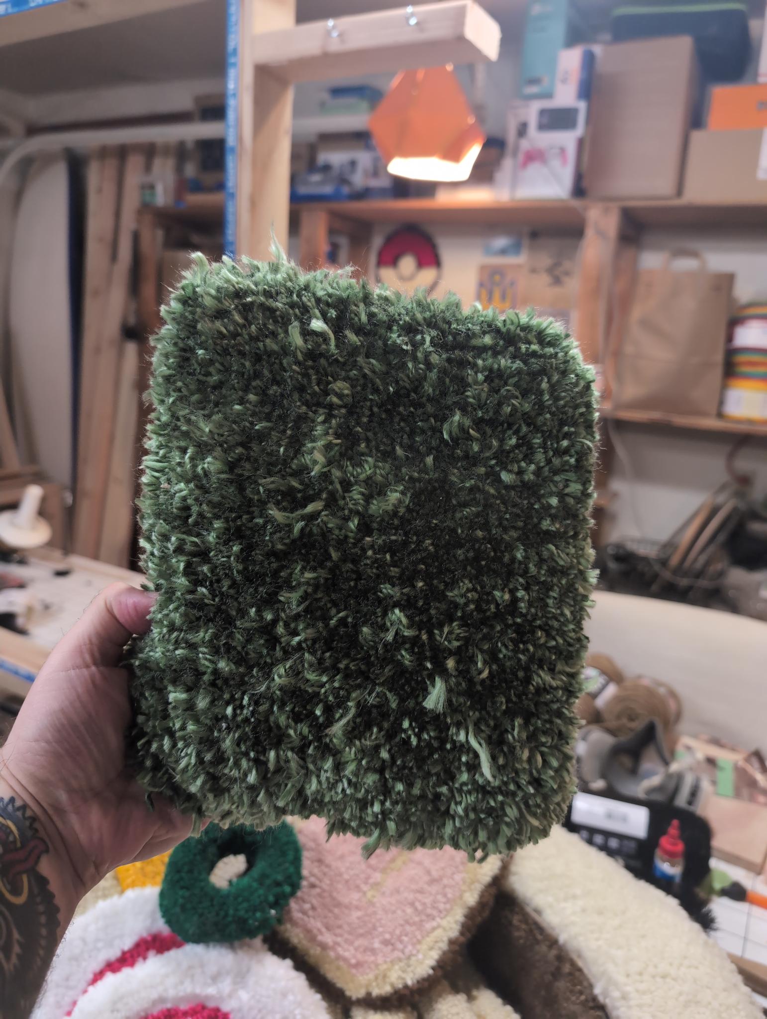 The tufted nori held in one hand, dense dark green shaggy pile, filling the whole hand and then some